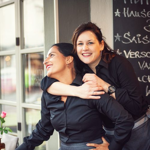 Zwei lachende Frauen, eine wird von anderer umarmt, tragen Arbeitskleidung für Gastronomie