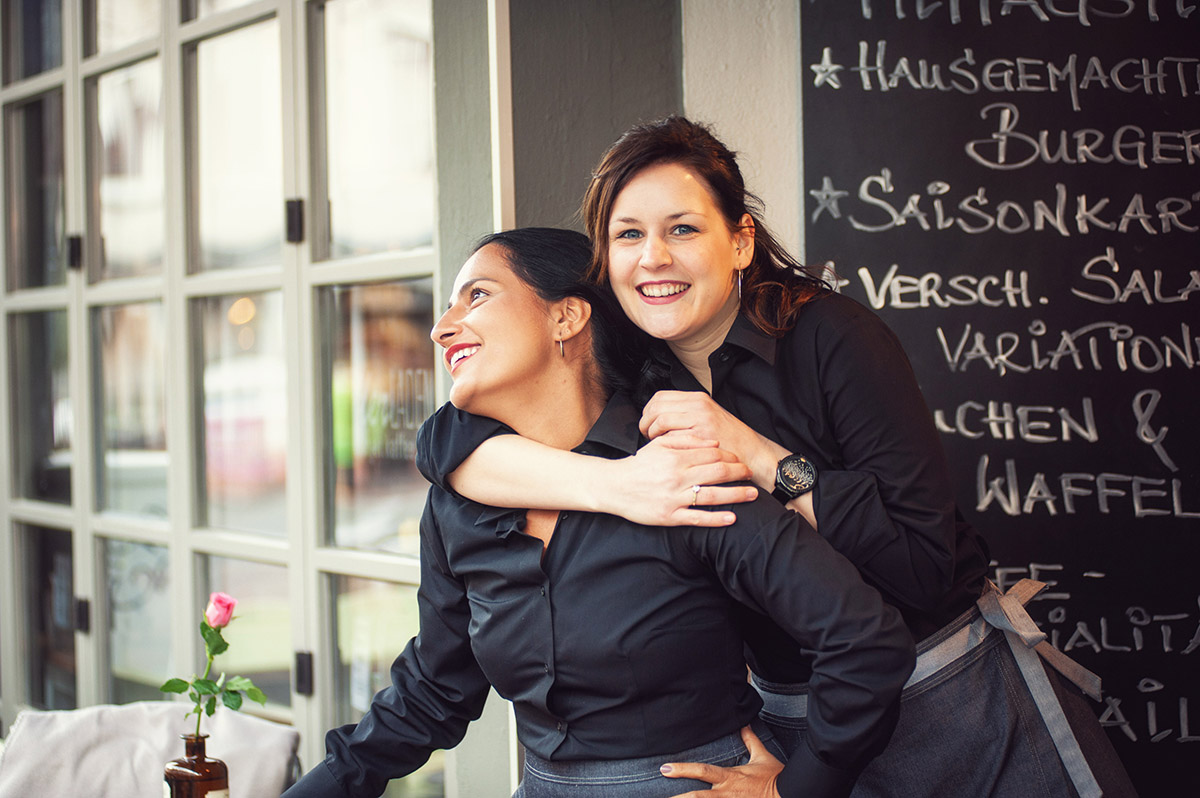 Zwei lachende Frauen, eine wird von anderer umarmt, tragen Arbeitskleidung für Gastronomie
