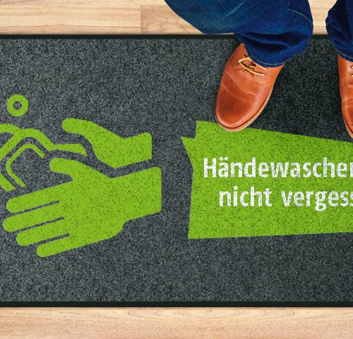 Mann mit blauer Jeans und braunen Lederschuhen steht auf Matte; Text: Händewaschen nicht vergessen!