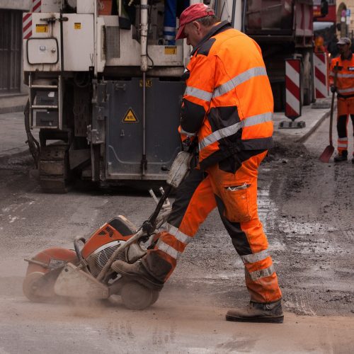 Arbeiter im Straßenbau, die Warnschutzkeidung tragen, im Vordergrund eine Person, die Teersäge bedient