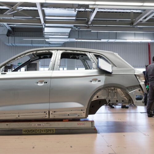Halle der Automobilindustrie, Autokarossiere aufgebockt, mehrere Personen stehen in Arbeitskleidung daneben