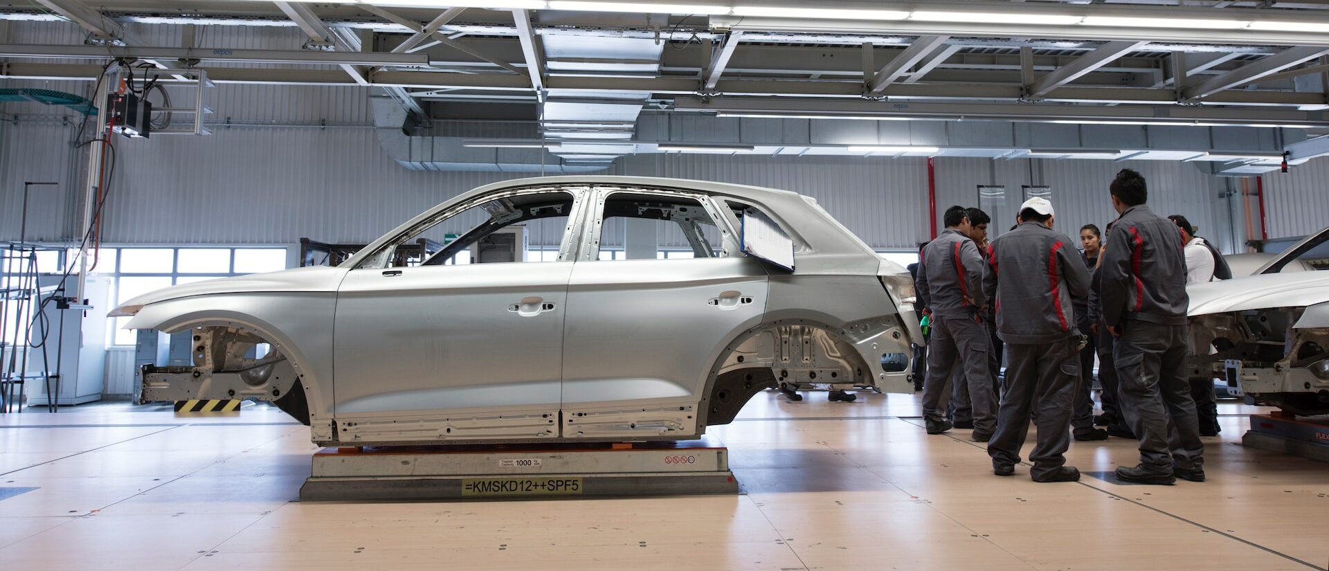 Halle der Automobilindustrie, Autokarossiere aufgebockt, mehrere Personen stehen in Arbeitskleidung daneben