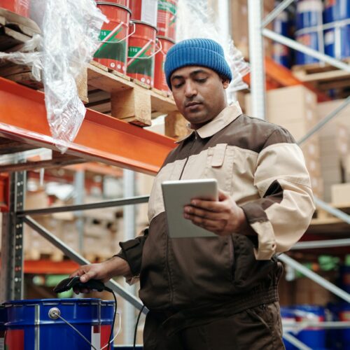 Ein Lagermitarbeiter in einer Mütze und Arbeitskleidung überprüft mit einem Tablet und Barcode-Scanner die Bestände in einem gut organisierten Lagerhaus